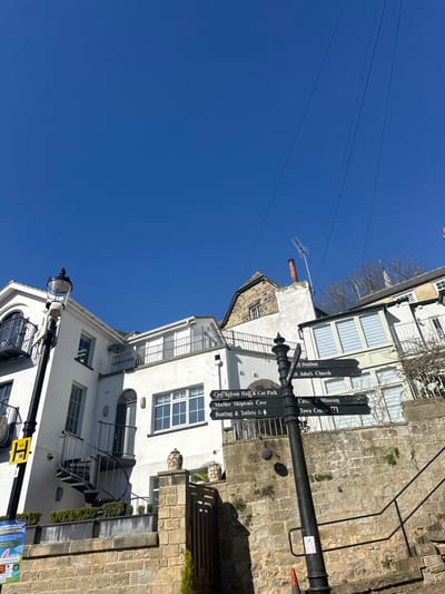 This image captures a daytime scene in Knaresborough, United Kingdom, under a clear, deep blue sky with no clouds. The perspective is looking uphill, with buildings constructed into the hillside.

In the immediate foreground, a low, tan-colored stone wall runs across the bottom. On the far left, partially obscured, is a blue and white information poster. To its right stands a black lamppost with an ornate top, featuring a yellow square sign with a black "H" (likely a bus stop sign) attached to its pole. Further to the right and slightly elevated is a black metal directional signpost with multiple arms. These arms, from bottom to top, point towards: "Boating & Toilets", "Mother Shipton's Cave", "Conyngham Hall & Car Park" (all pointing left/downhill), and "Town Centre", "Museum", "Castle", "St. John's Church", "Bus Station" (all pointing right/uphill). The base of the signpost is near a small orange traffic cone. A small evergreen plant is visible near the base of the stone wall.

Immediately behind the directional signpost and to its left, a light tan stone wall continues upwards, topped by a brown wooden gate and two decorative, patterned urns on stone pillars. To the right of the signpost, a black metal handrail with stone steps ascends the incline, though the steps themselves are mostly out of frame.

Several buildings populate the midground, primarily painted white with various architectural features. On the left, a multi-story white building features black wrought-iron balconies on its upper levels and a prominent external black spiral staircase leading to a doorway. To its right, another white building is visible, also with a balcony. Higher up and centrally located is an older building with exposed stone elements on its upper section, a red-brick chimney, and a television antenna on its roof. Further to the right, another light-colored building with multiple large, dark-framed windows is visible.

Overhead, multiple thin, dark wires (likely utility lines) stretch across the clear sky from the upper left to the upper right. Sparse, bare tree branches are visible against the sky on the upper right, and a distant, light-colored spire is faintly visible behind the buildings on the right. No people or animals are present in the scene. The lighting indicates direct sunlight, casting shadows to the left, suggesting the sun is generally to the right of the frame.