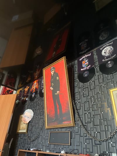 The image depicts a wall adorned with framed album covers and a large portrait, suggesting a room dedicated to music memorabilia. Prominently displayed is a portrait of a man in a military-style uniform, possibly Lemmy Kilmister from the band Motörhead, given the presence of several Motörhead album covers. The albums, some with vinyl records attached, are arranged in a grid-like fashion. A cut-out of a person's face, appearing to be Johnny Depp, hangs from a chain, adding a quirky element to the display. The wall itself has a distinctive dark grey patterned wallpaper. In the lower portion of the image, a wooden shelf unit is visible, containing some electronic devices and cables. The lighting suggests an indoor environment, possibly a living space or a collector's room. No specific location in Ishimbay, Russia is identifiable from the visual information in the image.