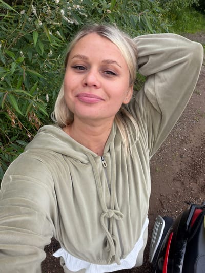 A young Caucasian woman with blonde hair pulled back and styled with a middle part is taking a selfie outdoors. She is wearing a light green velour tracksuit, which is zipped up partially, with a white textured fabric visible underneath at the hem. Her right arm is bent at the elbow and raised behind her head, with her hand resting on her scalp. She has light makeup on, with noticeable eyeliner and lip gloss. The background consists of lush green foliage on the left, and a dirt path on the right, possibly indicating a rural or park-like setting. Part of a black and red motorcycle or scooter is visible in the bottom right corner of the frame. The woman's expression is a slight smile, and she appears to be in good spirits. The lighting suggests it is daytime. There is no information in the image to suggest the location is in the city of Yaroslavl, Russia.