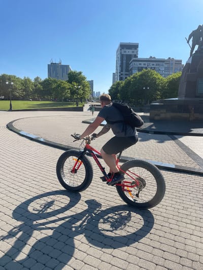 In Armyanskii, Russia, on a sunny day, a man with tattoos on his arms is riding a red fat-tire bicycle on a paved walkway. He is wearing a dark t-shirt, dark shorts, and dark athletic shoes, with a dark backpack slung over his shoulders. The walkway is made of light-colored rectangular pavers arranged in a circular pattern. To the left of the walkway is a large grassy area with scattered trees and a few lampposts. In the background, there are several modern apartment buildings and a large monument. The man's shadow and the shadow of his bicycle are cast prominently on the paved surface due to the bright sunlight.