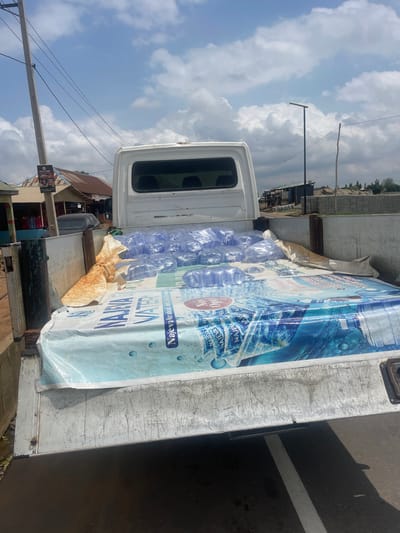 The image shows a white truck carrying a load of bottled water. The truck is parked on a dirt road next to a building. The sky is blue with some white clouds. The bottles of water are arranged in rows and are wrapped in plastic. A large banner with the words "NAJEVA WATER" is draped over the water bottles. The banner has images of water droplets and waves. The truck appears to be in a rural or semi-urban area, possibly in Tasha, Nigeria, given the presence of traditional-style buildings and overhead wires. The lighting suggests it is daytime, likely in the morning or afternoon. The scene seems to be a commercial delivery or transport of goods. There is no visible activity of people, only the parked truck and its cargo.