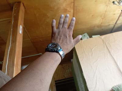 In Churapcha, Russia, a person's left arm and hand are visible, raised and facing the camera. The hand is open with fingers extended. A dark-colored digital watch with a metallic band is worn on the wrist. The arm and hand appear to have a light skin tone with some minor marks. The background consists of wooden paneling on the ceiling and walls, with some boxes and fabric visible in the distance. The lighting suggests an indoor setting.