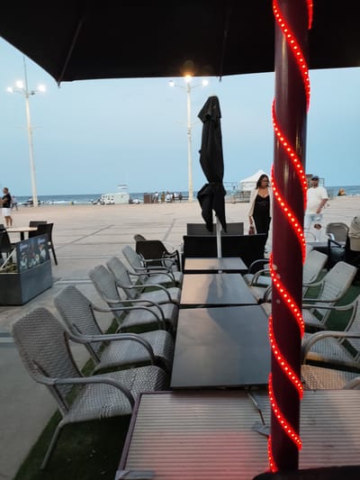 This image depicts an outdoor dining area at dusk, likely in Canet-en-Roussillon, France, given the coastal setting. The foreground is dominated by a patio with several tables and chairs. The chairs are of a light, woven material with metal frames. A dark, closed umbrella is visible in the middle ground, along with more tables and chairs. A dark red pole wrapped in a spiral of red LED lights stands prominently to the right of the frame.

In the background, people are seen walking and sitting on a sandy beach adjacent to a paved promenade. The sea is visible behind them, with a clear, pale blue sky above. A white building, possibly a lifeguard station or beach facility, is situated on the beach. A few streetlights with multiple bulbs are also visible, indicating a public area. Two individuals are walking towards the right side of the frame in the middle ground: a woman in a black outfit and a cream-colored jacket, and a man in a white t-shirt and distressed jeans. The overall atmosphere suggests a relaxed evening scene by the coast.