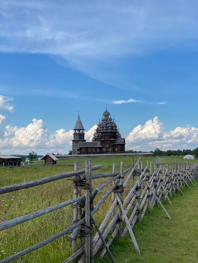 In Boyarshchina, Russia, a historic wooden church with multiple domed roofs and a bell tower stands prominently in a green, grassy field under a blue sky with scattered white clouds. A rustic wooden fence made of logs and branches runs diagonally across the foreground, separating the viewer from the church and a smaller wooden structure to its left. In the background, a body of water is visible, with trees and more distant structures lining the horizon. The scene is tranquil and appears to be a preserved historical site.