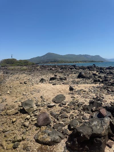 The image depicts a rocky beach with sand and sparse vegetation under a clear blue sky. In the distance, a chain of mountains forms the horizon, meeting a calm blue sea. The foreground is dominated by dark, volcanic rocks of various sizes scattered across the sandy terrain. A small island with green bushes and a flag pole is visible to the left. A solitary figure is seated on a rock near the water's edge. The scene is set in Lichada, Greece, known for its volcanic islands and rugged coastline.