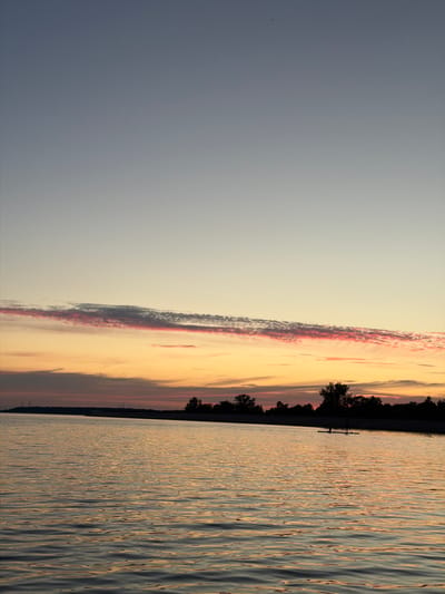 The image depicts a serene sunset over a body of water, likely the Daugava River in Salaspils, Latvia, given the context. The sky transitions from a soft pale blue at the top to hues of orange and pink near the horizon, with a distinct band of pink-tinged clouds stretching horizontally. The water's surface reflects the colors of the sky, creating ripples of light and shadow. Along the distant shoreline, a dark silhouette of trees and vegetation is visible, punctuated by what appear to be two individuals on paddleboards. The overall atmosphere is tranquil and evokes the end of a day.