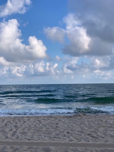 This is an image of a beach scene. The foreground consists of a sandy beach with visible footprints and ripples in the sand. The middle ground is occupied by the ocean, with waves gently breaking on the shore. The water is a deep blue-green color. The background is a sky filled with a mix of white and gray clouds against a clear blue sky. The scene depicts a calm day at the coast. The location is not specified in the image, therefore it cannot be identified as Rewal, Poland.