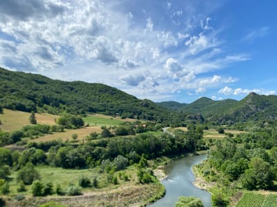 The image depicts a scenic landscape in Raska Municipality, Serbia, characterized by rolling green hills, a winding river, and a vast blue sky dotted with fluffy white clouds. The foreground features the Uvac River, its calm waters reflecting the surrounding greenery. The riverbanks are lined with lush trees and vegetation.

Further inland, the terrain rises into gently sloping hills covered in dense forests. Patches of open fields, some with cultivated crops, are interspersed among the trees, adding a touch of rural charm. A winding road is visible in the mid-ground, snaking its way through the landscape, hinting at human presence and connectivity within this natural setting. Small houses or farm buildings are sparsely scattered throughout the scene, blending into the environment.

The sky above is a brilliant blue, with scattered cumulus clouds that create dynamic patterns of light and shadow across the hills. The overall impression is one of serene natural beauty, typical of the Serbian countryside. There are no people or animals visible in the image.