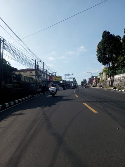 The image depicts an asphalt road in Nhot Chang, Laos, under a clear sky with a few clouds. The road is marked with a broken yellow line down the center. Power lines run across the top of the frame, appearing dense on the left and thinning toward the right. Buildings line the left side of the road, interspersed with trees and vegetation. A concrete wall topped with vegetation borders the road on the right. A person is riding a motorcycle down the road. The photo is taken from a low perspective, suggesting the viewpoint of someone on or near the road surface.