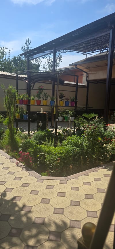 The image depicts a paved outdoor patio area with a garden in the background, situated in Uychi, Uzbekistan. The patio is paved with hexagonal and square tiles in a beige and light brown color scheme, featuring intricate star-like patterns on the hexagonal tiles. A decorative border runs along the edge of the patio.
In the background, a lush garden is visible, filled with various green plants, bushes, and some flowering plants with pink and red blossoms. A metal shelving unit stands in the garden, holding several colorful flower pots in red, yellow, blue, and pink. Some of these pots contain green plants. Below these, on a lower shelf, are more plants in white and green pots. A dark, rectangular structure with a white faucet is positioned in front of the shelving unit.
Overhead, a dark metal pergola or canopy structure is present, with decorative metal scrollwork. Beyond the garden and pergola, parts of a building with a light-colored wall and a tiled roof are visible, suggesting a residential or commercial property. The sky above is blue with scattered white clouds. The overall scene suggests a well-maintained outdoor space, likely a garden or patio area of a home or establishment. No people are visible in the image.