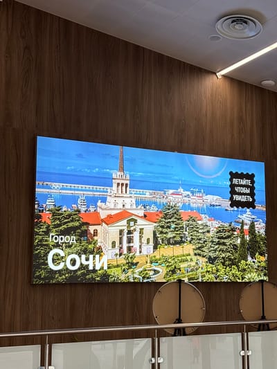 The image shows a vertical, large-format digital display mounted on a wooden wall. The display is showing a vibrant image of a coastal city with a clear blue sky and calm water. In the foreground of the display, there is lush green foliage and buildings with red roofs. A prominent architectural feature, resembling a tall tower or spire, is visible on the left side of the display. The sea is filled with yachts and boats docked along a waterfront. The Olympic rings symbol is visible in the water.

On the upper left portion of the display, there is a black stamp-like graphic containing white text in Russian. The text reads "ЛЕТАЙТЕ, ЧТОБЫ УВИДЕТЬ" which translates to "FLY TO SEE". Towards the bottom of the display, larger white text in Russian reads "Город Сочи" meaning "City of Sochi".

The overall scene depicted on the screen suggests a sunny day, likely during warmer seasons, promoting tourism or a sense of travel. The lighting on the wooden wall and the ambient light suggest the photograph was taken indoors, possibly in a lobby or public space. To the left of the display, a white circular ventilation grille is visible on a light-colored tiled surface. To the right of the display, metallic poles and glass panels suggest the presence of a modern interior design, possibly a hallway or an entrance area. The image is captured from a slightly upward-angled perspective, emphasizing the size and placement of the digital display.