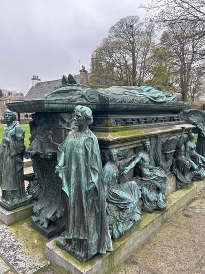 The image shows a close-up, tilted view of a bronze monument, located in Aberdeen City, United Kingdom. The monument features multiple figures, likely representing a historical or allegorical scene. The figures are draped in clothing and possess intricate detailing. The bronze has a green patina, suggesting age and weathering. The monument rests on a stone base, which is covered in moss and lichen. In the background, bare trees and a building with a grey roof and chimneys are visible. The sky appears overcast.