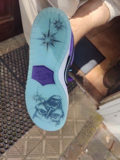 A person is holding up a sneaker to display its sole. The sneaker features a light blue translucent sole with a dark purple Nike SB logo. The sole is scuffed with what appears to be black residue. The upper part of the shoe is visible, with purple and white accents. The location appears to be a doorway, with a doormat composed of raised honeycomb-shaped patterns. A brown tiled surface and a black welcome mat are visible inside the doorway. The image does not provide enough context to definitively state the location is in Dorst, Netherlands.