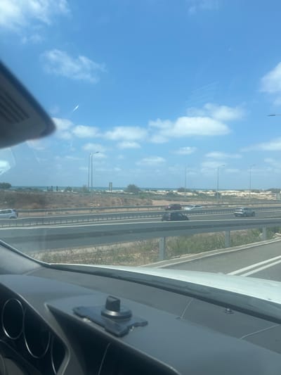 The image captures a view from inside a vehicle, looking out at a highway. The foreground shows a portion of the car's dashboard and windshield. The highway itself is visible in the midground, with several cars traveling on it. Beyond the highway, there is a sandy landscape leading to the sea, which is visible in the distance. The sky is blue with scattered white clouds. There are also streetlights visible along the highway. The location is identified as Hofit, Israel. The overall scene depicts a typical roadside view on a clear day.