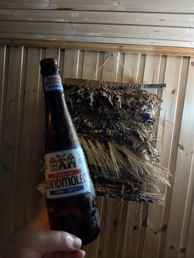 A hand holds a partially visible brown glass beer bottle in front of a wooden wall. The bottle has a label with red and blue accents and features an illustration of houses and a windmill, along with text that reads "BLANCHE" and "WINDMÖLER". Behind the bottle, a decorative arrangement of dried plants and grasses hangs on the wall, also constructed from natural materials. The wall is made of vertical wooden planks with visible knots and grain. The image appears to be taken indoors, possibly in a rustic setting. No specific location in Nizhnetroitsky, Russia is evident from the image itself.