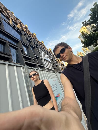 A low-angle, wide-angle photograph captures a man and a woman posing outdoors on a sunny day in what appears to be an urban environment, specifically Putuo District, China.

The man, positioned on the left side of the frame, wears a black t-shirt and dark sunglasses. He has dark hair and a neutral expression as he looks directly towards the camera. His left arm, visible in the foreground, shows some hair.

The woman, on the right side of the frame, wears a black tank top and dark sunglasses. She also looks directly at the camera with a neutral expression. Her right arm and hand are visible in the lower right foreground.

Behind the individuals, a modern, multi-story building with a distinctive geometric facade dominates the left side of the scene. It features dark glass windows and angular architectural elements, with some greenery visible on its upper levels. A temporary, corrugated metal fence or barrier runs horizontally across the lower middle of the frame, separating the subjects from the building in the foreground.

The sky, bright blue with scattered wispy white clouds, fills the upper and right portions of the image, indicating clear, sunny weather. Other buildings and trees are faintly visible in the distant background.

An unreadable white sign with black Chinese characters is attached to the corrugated fence behind the woman, suggesting a public notice or informational board. The overall impression is a casual outdoor snapshot taken in bright daylight.