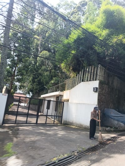A man sweeps debris from a paved road. He wears a light brown shirt, dark pants, and a baseball cap. Behind him is a white wall with the number "3" on it and a tall black metal fence. A small white building is next to the wall. Above the wall is a fence with tall metal bars and lush green foliage. Trees line the background, with numerous power lines strung above.