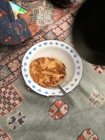 The image shows a close-up of a child's feet and legs wearing patterned pajamas, resting on a patterned blanket. A tablet is visible, displaying a colorful game. In the foreground, a round, golden-brown pancake sits in a white enamel bowl with blue trim. A silver fork rests on the pancake and extends out of the bowl. The blanket has a complex geometric pattern with shades of blue, orange, and tan. The scene appears to be indoors, with a dark, out-of-focus object in the upper right corner possibly being a piece of furniture or another person. There is no specific indication of the location being Sungai Meriam, Indonesia.