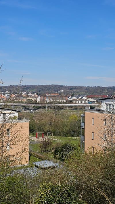 The image captures a view of Rheinfelden, Switzerland. A residential area is interspersed with green spaces and trees. Buildings vary in height and color, with some featuring modern architectural designs. An elevated metal structure spans the cityscape, possibly a railway or a bridge. The sky is a clear blue with some trees in the foreground. The image is oriented vertically.