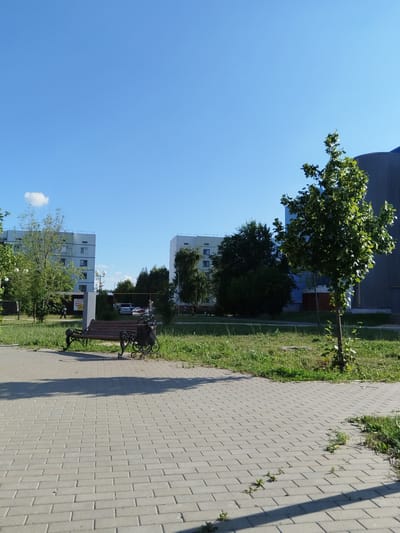 The image depicts an outdoor scene on a clear day, likely in the city of Roshchinsky, Russia. In the foreground, a grey brick pathway stretches across the bottom half of the frame. To the left and slightly behind the pathway, a dark-colored park bench with ornate metal legs and a trash receptacle are situated on a patch of green grass.

Beyond the bench, two multi-story apartment buildings with light-colored facades and numerous windows are visible. A few trees and dense foliage are scattered between the buildings and in the midground. Further to the right, a tall, slender building with a ribbed grey exterior, possibly a modern structure or industrial building, is partially visible, with a small, leafy green tree in front of it. The sky is a solid, bright blue with a single small white cloud. People are sparsely present, with a few individuals walking in the background near the apartment buildings and vehicles are parked in the distance. The overall atmosphere suggests a residential area during daytime.