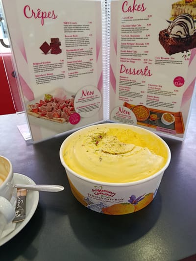 The image displays a menu from an establishment, likely a cafe or dessert shop. The menu is divided into sections titled "Crêpes," "Cakes," and "Desserts." Prominently featured in the foreground is a large tub of yellow ice cream, possibly Persian Saffron flavor, as indicated by the text on the tub. A cup of coffee and a sugar packet are visible on a saucer to the left of the ice cream. The background shows the menu board with various dessert descriptions and prices, including items like "M&M Crunch," "Belgian Chocolate," "Carrot Cake," and "Chocolate Chip Cookie Dough." The lighting suggests an indoor setting. The image does not provide any specific information about its location in Bedford, United Kingdom.