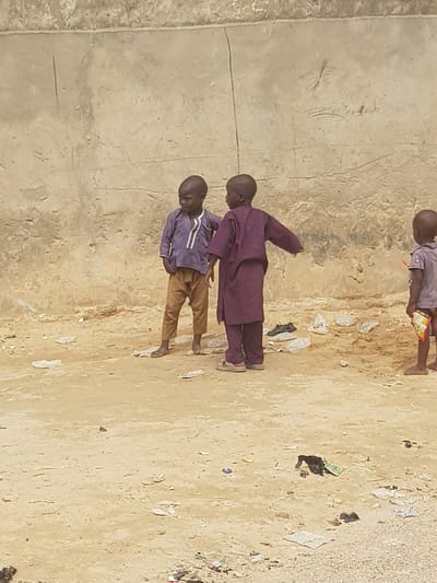 Three children are present in an outdoor setting in Dutsinma A, Nigeria. The ground is light brown and strewn with litter. Two children stand near a textured, light-colored wall. One child wears a purple outfit with khaki-colored pants. The second child wears a purple robe. The third child is in the background, and visible from the torso up, wearing a light-colored shirt and holding a food wrapper.