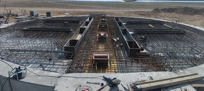 The image captures an aerial view of a construction site in Magnuszewice, Poland. The foreground features a large, circular concrete foundation reinforced with an intricate network of rebar. Several construction tools are visible on the concrete surface, including an angle grinder, a power cord reel, and various metal pieces. In the center of the rebar structure, a rectangular trench-like formwork is present, with some wooden supports inside. Beyond the foundation, the terrain transitions to bare earth and sand, with piles of construction materials, including lumber and scaffolding, scattered around. In the background, a vast, flat field is visible, possibly agricultural land, with large white structures, likely storage silos or temporary shelters, in the distance. The overall scene suggests ongoing construction work on a significant infrastructure project.
