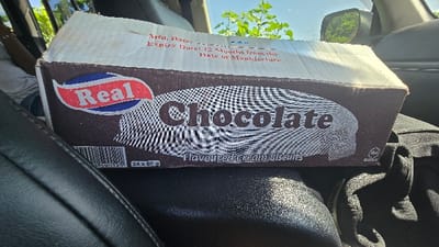An eye-level shot inside a vehicle in Shimbi Mashariki, Tanzania, features a large cardboard box of "Real Chocolate Flavoured Cream Biscuits" resting on a dark car seat. The box, predominantly brown with a white top, displays the brand name "Real" in red lettering within a blue and red oval logo on the left. The word "Chocolate" is prominently printed in large white letters across the center, above the description "Flavoured Cream Biscuits." Below the chocolate text, a textured, wavy pattern in black and white suggests the appearance of biscuits.
Visible text on the top flap of the box includes "Mfg. Date:" (Manufacturing Date) and "Expiry Date: 12 Months from the Date of Manufacture," though the actual manufacturing date is not clearly readable. On the lower left front, a barcode is partially visible, along with the quantity "24 x 60 g," indicating 24 individual biscuit packs, each weighing 60 grams. Towards the bottom right of the box, some faint text possibly indicates origin or certification, with "SRI LANKA" being partially discernible.
The car interior is visible, with dark leather or vinyl seats. A person's arm, wearing a bracelet, is partially seen on the left. Through the car window, bright daylight illuminates green foliage and a clear sky, suggesting a sunny day. A side-view mirror reflects green trees, further indicating the outdoor setting. The box appears to be in transit, possibly having just been purchased or on its way to a destination.