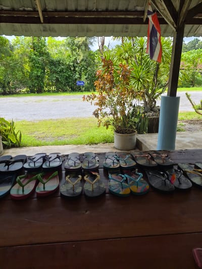 A row of colorful flip-flops are displayed on a wooden surface, likely for sale or for rent. Behind the flip-flops, a lush green landscape with trees and foliage is visible. A Thai flag hangs from the roof of an open-air structure, suggesting the location is in Thailand. A blue sign with Thai script is also visible in the background. The overall scene appears to be an outdoor setting, possibly at a beach resort or a market in Phang-nga, Thailand.
