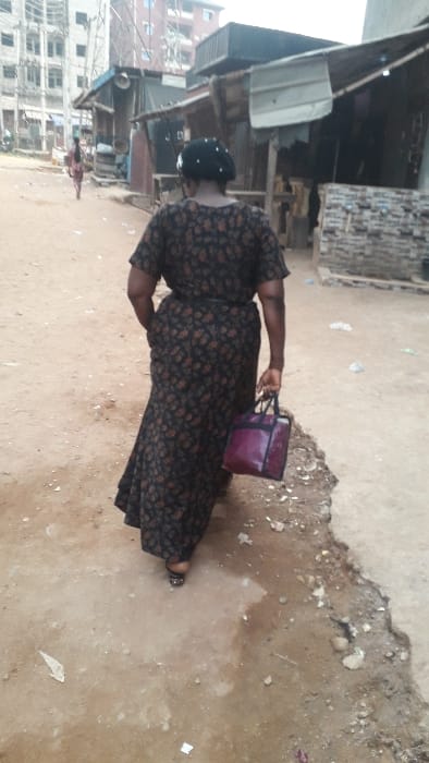 The image shows an outdoor scene in what appears to be a residential or semi-urban area in Awada, Nigeria. The primary subject is an adult, likely a woman, seen from behind and slightly to her left, walking on an unpaved path. She is wearing a long, dark-colored dress with a lighter, perhaps floral or abstract, pattern, and a dark head covering. In her right hand, she carries a burgundy or purple handbag with dark straps. Her left foot, visible near the bottom of the frame, is wearing a light-colored sandal or slipper.

The ground is uneven, composed of dirt and sand, with visible patches of dampness or mud, along with scattered small rocks and debris. To the left of the path, various structures are visible, including what appears to be wooden shacks or makeshift buildings. A white, rectangular object, possibly a large container or discarded appliance, sits near one of these structures. Further in the background on the left, a multi-story building under construction can be seen, indicating development in the area. The lighting suggests it is daytime, with diffused light rather than direct sunlight, possibly indicating an overcast sky. There is no visible text in the image. The scene conveys a glimpse into daily life on a typical pathway in a developing neighborhood.