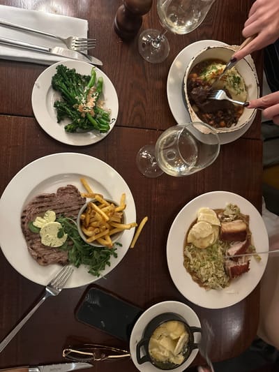 The image shows a table at a restaurant, likely in St Albans, UK, based on the style and types of dishes. There are at least four people dining, evident from the various plates of food and hands visible. Dishes include:
* A stew or casserole with mashed potatoes in a distressed white bowl.
* Pork belly with shredded cabbage and potatoes.
* Broccolini topped with nuts.
* A steak with fries served in a metal cup and arugula.
* A dish of sliced potatoes, possibly dauphinoise.
Each place setting has silverware. There are also glasses of white wine on the table. A smartphone and glasses case are placed on the table next to one of the plates. The table itself is a dark, polished wood. The people are in the process of eating, as forks are in motion and some food has already been consumed.