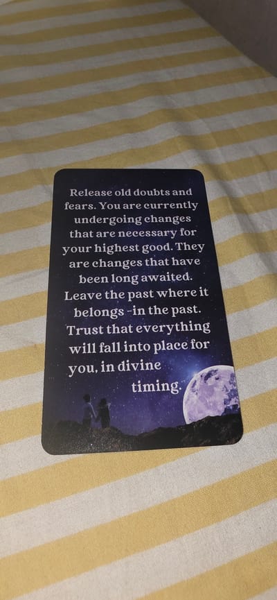 The image shows a close-up, top-down view of a dark blue card with white text and a starry night sky graphic. The card is lying on a yellow and white striped surface, possibly a tablecloth or bedding. The text on the card reads:
"Release old doubts and
fears. You are currently
undergoing changes
that are necessary for
your highest good. They
are changes that have
been long awaited.
Leave the past where it
belongs -in the past.
Trust that everything
will fall into place for
you, in divine
timing."
The graphic on the bottom half of the card depicts two silhouetted figures standing on a hill, holding hands, looking up at a large, luminous full moon and a starry sky.
The location in the city of Lieli, Switzerland is not determinable from the provided image.