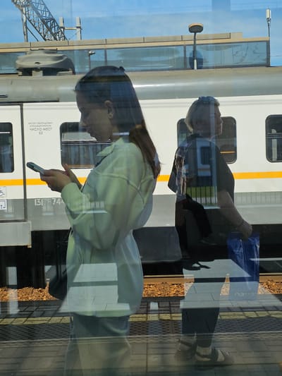 This is a medium-shot, eye-level, reflected image of two women standing on a train platform. The woman on the left is in the foreground, facing right, and is looking down at her phone, which she is holding in both hands. She has long brown hair and is wearing a light-colored jacket. The woman on the right is in the background, facing right, and is looking forward. She has short blonde hair and is wearing a dark t-shirt and dark pants. She is holding a blue shopping bag in her right hand.

Behind the women is a white train with a yellow stripe. The train is stopped at a platform. The platform has a yellow tactile warning strip along its edge. The ground on the platform is paved with light-colored tiles. There are train tracks with gravel ballast visible beyond the platform.

The image is a reflection on a glass surface, likely the window of a train or a building. The reflections of the women and the train are visible on the glass. The sky is visible through the glass, with some clouds. The location is identified as the Novoivanovskoe, Russia train station.