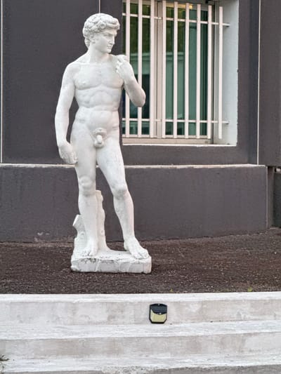 In the city of Grenay, France, a white marble statue resembling the classical sculpture of David stands outdoors. The statue depicts a nude male figure with curly hair, in a contrapposto stance, with his right hand resting on his right shoulder. He stands on a rectangular base. Behind the statue, a dark gray building with a white-trimmed window featuring vertical bars is visible. The ground around the statue is covered with dark gravel, and a set of concrete steps leads down from the foreground. A small, black and yellow outdoor light fixture is positioned on the second step from the bottom.