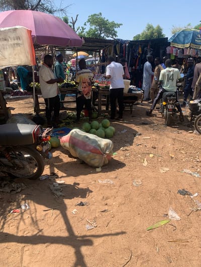 This media file captures a bustling outdoor market scene in Nigeria. The image is oriented vertically, presenting a ground-level view of a busy marketplace. Several individuals are visible, appearing to be vendors and shoppers. One person in the foreground is wearing a white shirt and black pants, and another to their left wears a white shirt with black sleeves. In the background, more people are present, some interacting with market stalls laden with goods.
The market is set up with wooden structures and colorful umbrellas, offering shade. Various items are displayed for sale, including clothing hanging from a stall under a blue umbrella with "MoM" text, and a pile of produce, likely watermelons, near a parked motorcycle. The ground is dusty, characteristic of many outdoor markets. Scattered on the ground are pieces of litter, including plastic bags and other debris, suggesting a well-used, unpaved area.
A motorcycle is parked prominently in the middle right of the frame. There's a large, roughly woven sack filled with what appears to be produce, possibly watermelons, next to it. The sunlight suggests it is daytime, likely late morning or afternoon, casting distinct shadows on the dusty ground. The overall atmosphere appears lively and active, typical of a Nigerian market.
Visible text includes "MoM" on an umbrella, "Dial *671#" with instructions, "ALE & PLLHJTUFHCO" and "FLOW" on clothing, "SUNSI" on a shirt, and the number "14" on a light green shirt. The "CH" on the motorcycle's license plate is also visible.
