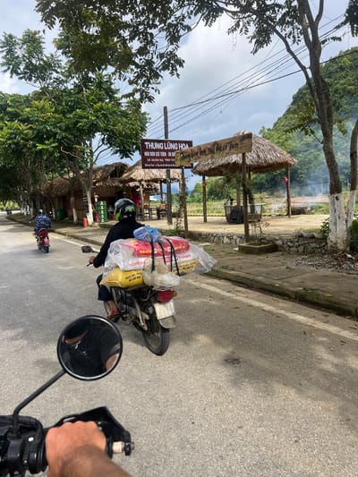 In Quản Bạ Commune, Vietnam, a motorbike rider, wearing a black jacket and helmet, transports several large sacks of what appears to be grain or feed, secured on the back of their motorcycle. The sacks are yellow and red, and one prominently displays the text "RED STAR". The rider is in the foreground, viewed from behind. Ahead, another motorbike rider, wearing a camouflage patterned jacket, is visible on the road. The scene is set along a paved road lined with trees and traditional thatched-roof structures. A sign above one of the structures reads "THUNG LŨNG HOA NÚI ĐÔI CỔ TÍCH" which translates to "Fairytale Twin Mountain Valley". In the background, lush green hills and a cloudy sky complete the landscape.