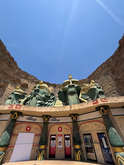 The image shows an exterior view of the Coca-Cola Freestyle station at Gardaland Park, located in Castelnuovo del Garda, Italy. The facade is designed in an Egyptian theme, featuring large, stylized green and gold heads resembling pharaohs or gods, carved into a rocky cliff-like structure. Below these heads, a decorative ledge is supported by ornate pillars, also in a green and gold palette with Egyptian motifs. The area features several Coca-Cola Freestyle machines, some with interactive screens, and signage. A blank white panel is leaning against the wall to the left. The sky above is clear blue with faint contrails from an aircraft. The ground appears to be paved.