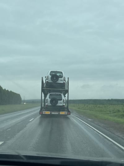 A flatbed trailer carrying two white SUVs is seen from the perspective of a driver on a wet, two-lane highway. The trailer is positioned to the right of the frame, with one SUV on the lower deck and another on the upper deck. Both vehicles have spare tires mounted on their rear doors. The lower SUV has a license plate that reads "ev 0414 50". The highway is surrounded by lush green fields and a line of trees on the left side. The sky is overcast and the road surface is wet, indicating it is raining or has recently rained. The location is Arkhangelskoye, Russia.