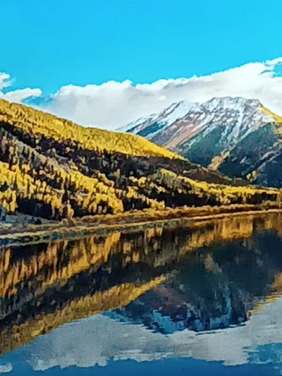The landscape showcases a placid lake reflecting mountains under a bright blue sky with scattered clouds. A range slopes down to the water's edge, covered in dense, yellow-leaved trees. A snow-capped mountain rises in the background. The reflection in the water mirrors the scene above, creating a symmetrical composition.