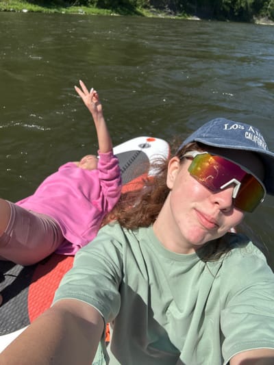 In a close-up, overhead selfie taken on a paddleboard, two young females are partially visible in the foreground and midground against a backdrop of dark green water and a tree-lined bank. The female in the foreground, wearing a light green crewneck t-shirt and a blue baseball cap with "LOS ANGELES CALIFORNIA" embroidered on it, smiles at the camera while sporting large, multicolored mirrored sunglasses. Her right arm, extended towards the viewer, is a selfie arm, capturing the scene. In the midground, another female, lying down on the paddleboard, wears a bright pink hooded sweatshirt and light-colored shorts. Her left arm is raised, with her fingers forming a peace sign, and her head is tilted back. The paddleboard, featuring a black and red patterned deck, is partially submerged in the rippling water. The overall setting suggests an outdoor recreational activity on a body of water, likely during daylight hours. The location is not Manskii, Russia.
