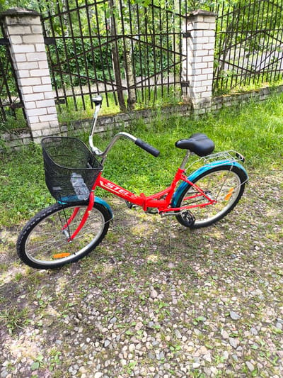A red and blue bicycle with a black basket is parked on a gravelly path next to a green, grassy area. The bicycle is a step-through frame model with a black seat and handlebars, a silver rack over the rear wheel, and a black wire basket on the front. The word "STEL" is visible in white lettering on the red frame. In the background, a metal fence with white brick posts borders the grassy area. The scene appears to be outdoors on a sunny day, with lush green foliage visible through the fence. The location is identified as Novonikolskoye, Russia.