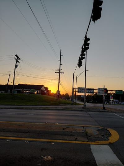 This is a photograph of an intersection at sunrise in Frankfort, Kentucky, United States of America. The sky is a pale orange and yellow, indicating the early morning sun. Several telephone poles and power lines stretch across the sky, silhouetted against the sunrise. In the background, a long, low building with a dark roof sits on a grassy hill. A sign with green lettering can be seen in front of the building. To the right of the frame, a traffic light hangs over the intersection. Further down the road, highway signs indicate "460 East Georgetown." A gas station with a Marathon Food Mart sign is also visible. The foreground shows an asphalt road with yellow lane markings and a crosswalk. The overall scene depicts a quiet, early morning at a suburban intersection.