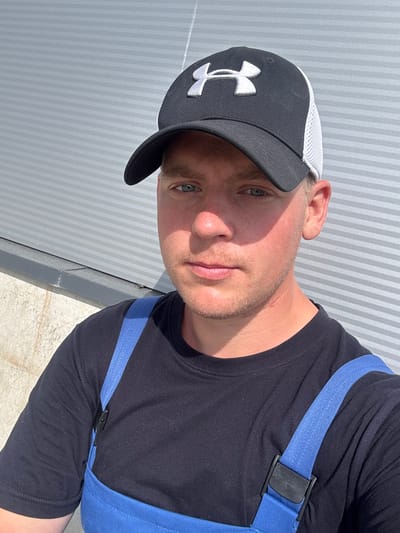 This is a close-up, rotated image of a person wearing a black t-shirt and a black baseball cap with a white mesh back. The cap has a white Under Armour logo on the front. Blue straps are visible over the person's shoulders, attached to a black clip. The person has light-colored eyes and stubble. The background consists of a corrugated metal wall with a faint striped pattern, suggesting an outdoor setting in daylight. The lighting indicates it is daytime, likely sunny, casting a shadow on the person's face. The setting appears to be Sauleskalns, Latvia. No specific activity is clearly depicted, but the attire and straps might suggest work or a specific activity requiring them.