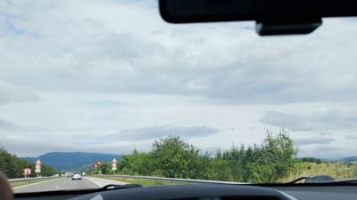 The image is a point-of-view shot from inside a vehicle traveling on a highway in Tserovo, Bulgaria. The road ahead is visible, with multiple lanes and a white dividing line. Several cars are on the road. On the right side of the road, there is a metal guardrail and a patch of tall grass and trees. On the left side, there are more trees, a guardrail, and several traffic signs, including a speed limit sign indicating 120. In the distance, there are rolling green hills and mountains under a cloudy sky. The interior of the car is partially visible at the bottom of the frame, including the dashboard and part of the windshield.
