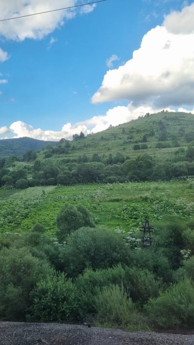 This is a landscape photograph taken in Yasenytsya-Zamkova, Ukraine. The image features a rolling, green hillside covered in trees and vegetation. The sky is blue with scattered white clouds. In the foreground, there is a dark, rough surface, possibly gravel or asphalt. A wooden utility pole with insulators is visible on the hillside.