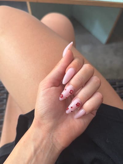 A close-up, eye-level shot shows a person's left hand with light pink almond-shaped fingernails, adorned with small red rhinestones arranged in a floral pattern. The hand rests on black fabric, with the person's bare leg visible in the background. The environment appears to be outdoors, possibly a cafe or patio, with out-of-focus wooden furniture and a teal-colored wall in the distance. The scene is lit by natural light, casting soft shadows. The location is not discernible from the image.