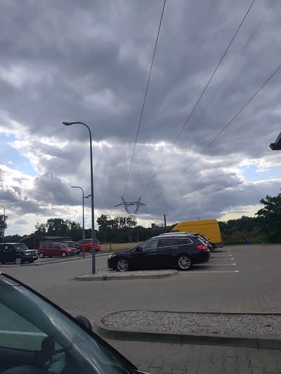The image depicts a parking lot with several cars, a yellow van, and two lampposts under a cloudy sky. In the foreground, the reflection of a car windshield is visible. In the midground, a black station wagon is parked, facing right. Behind it, a yellow van is parked, also facing right. Further back, red and black cars are parked. The background consists of trees and a power transmission tower. The scene appears to be located in the city of Czapury, Poland, as indicated by the presence of typical European license plates on some vehicles. The overall atmosphere is overcast, with sunlight breaking through some of the clouds.