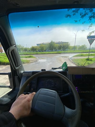 The image captures a first-person perspective from inside a vehicle, likely a truck or van, looking out through the windshield. A driver's right hand rests on the steering wheel, indicating preparation for or a pause during driving. The dashboard is visible, featuring a speedometer with markings from 0 to 160, and a small tag with "TRW" visible near the windshield. A green air freshener or accessory labeled "SmellCar" hangs from the rearview mirror.
Outside the vehicle, a damp, unpaved road or dirt track is visible, with numerous puddles suggesting recent rain. Green grass and scattered trees line the sides of the road. In the mid-ground, several low-rise buildings with industrial or residential characteristics are present. The sky appears bright but overcast or hazy, with a blue tint on the upper part of the windshield. A triangular road sign with a red border and white background is partially visible above the trees. Power lines and utility poles are also seen in the distance. The setting suggests an urban fringe or industrial area within Usolye-Sibirskoye, Russia, during daytime under possibly cloudy weather conditions.