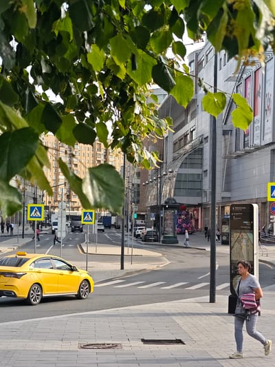 The image depicts a street scene in Moscow, Russia. In the foreground, a woman with dark hair, wearing a grey t-shirt and grey pants, walks on a paved sidewalk, carrying a striped bag over her shoulder. To her left, a yellow taxi cab is driving across a crosswalk. Behind the taxi, several cars and trucks are visible on the street. Pedestrian crossing signs are present on poles along the street. In the background, a large, modern building with glass and grey facades stands, adorned with multiple billboards. Further in the background, a tall apartment building with numerous windows is visible. Green foliage from trees hangs over the scene, partially obscuring the view of the buildings and sky. The overall atmosphere suggests an urban environment during daylight hours.