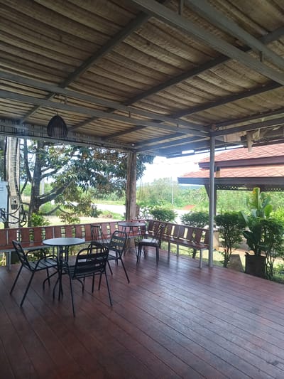 The image depicts an outdoor patio area, possibly a cafe or restaurant, in Plai Phraya, Thailand. The patio is covered by a corrugated roof supported by a metal frame. The floor is made of wood planks, stained a reddish-brown color.

The patio is furnished with several black metal tables and chairs. There are also built-in wooden benches along the perimeter. Plants and greenery border the edge of the patio on one side.

Outside the patio, there are trees and foliage, suggesting a rural or suburban setting. A road is visible in the background. A red-tiled roof structure is also visible nearby. The lighting suggests it is daytime. No people are visible in the image.